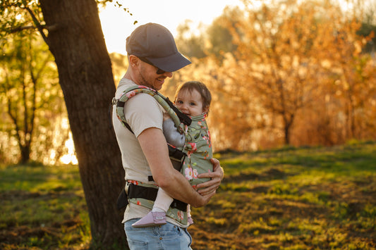 A father carrying his baby in a floral baby carrier while walking outdoors in an autumn park.