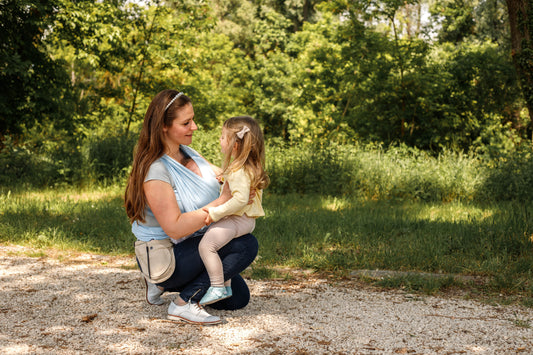 A mother wearing a light blue baby wrap holds her older daughter on her knee, while a younger baby rests in the wrap.