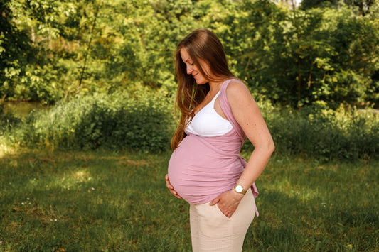 A pregnant woman wearing a pink baby wrap stands outdoors in a green park, gently holding her belly.