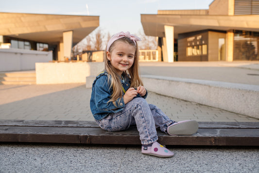 A young girl wearing pink soft-soled shoes sits on a wooden bench outdoors.