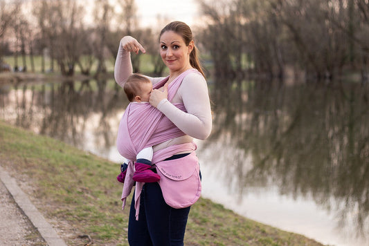 A mother breastfeeding her baby in a pink baby wrap while walking outdoors beside a pond.