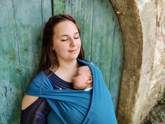 A mother peacefully babywearing her newborn in a blue wrap against a rustic turquoise wooden door.