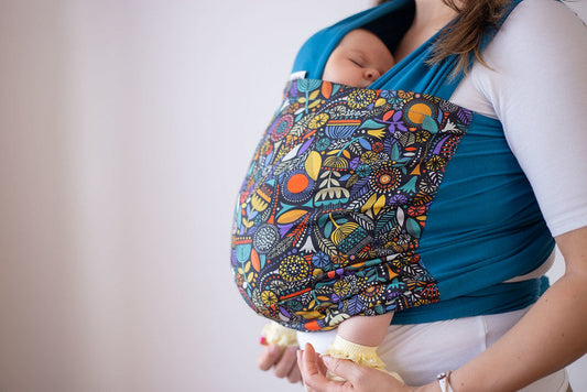 A sleeping baby rests in a colorful patterned baby carrier while being carried by their mother.